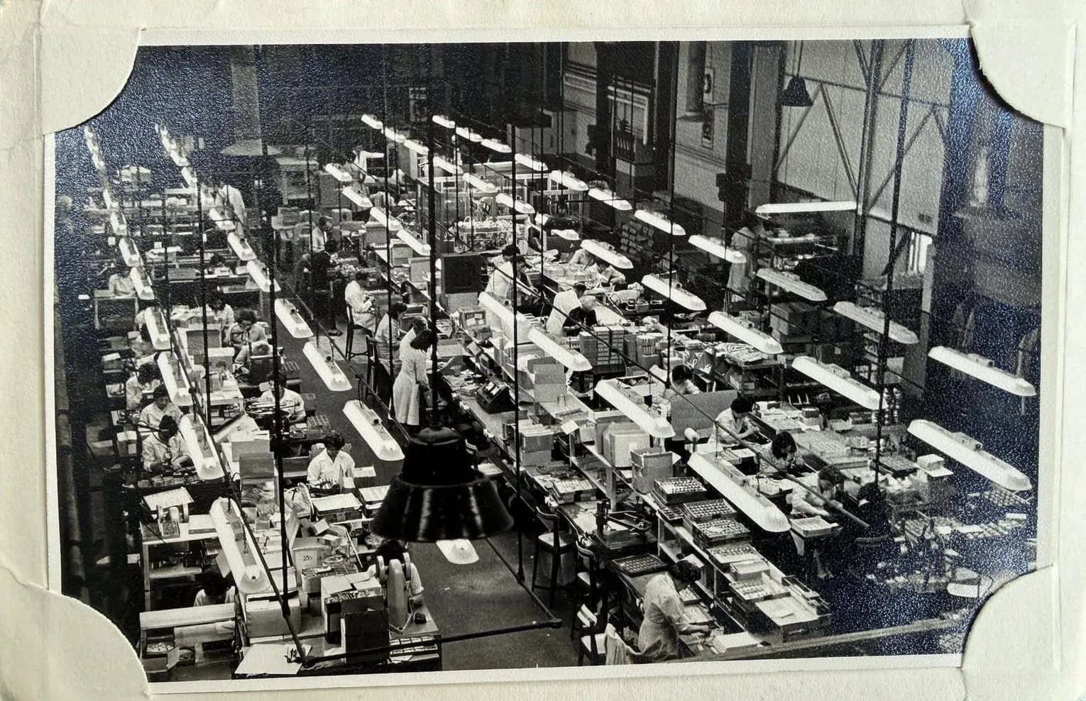 Black-and-white photograph, mounted in an album, of rows of workers at assembly benches under strip lights in the Ronson factory, seen from above.