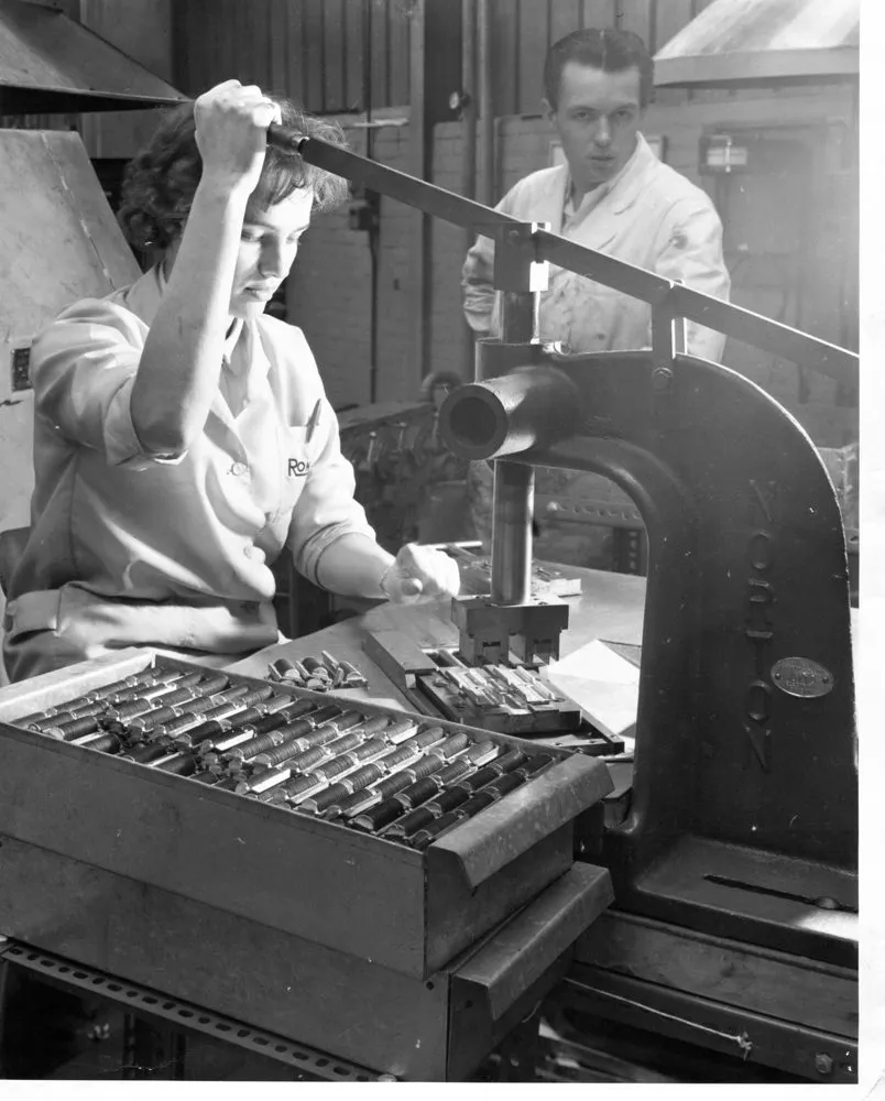 Black-and-white photograph of a woman operating a hand press at the Ronson factory, with a manager watching in the background.