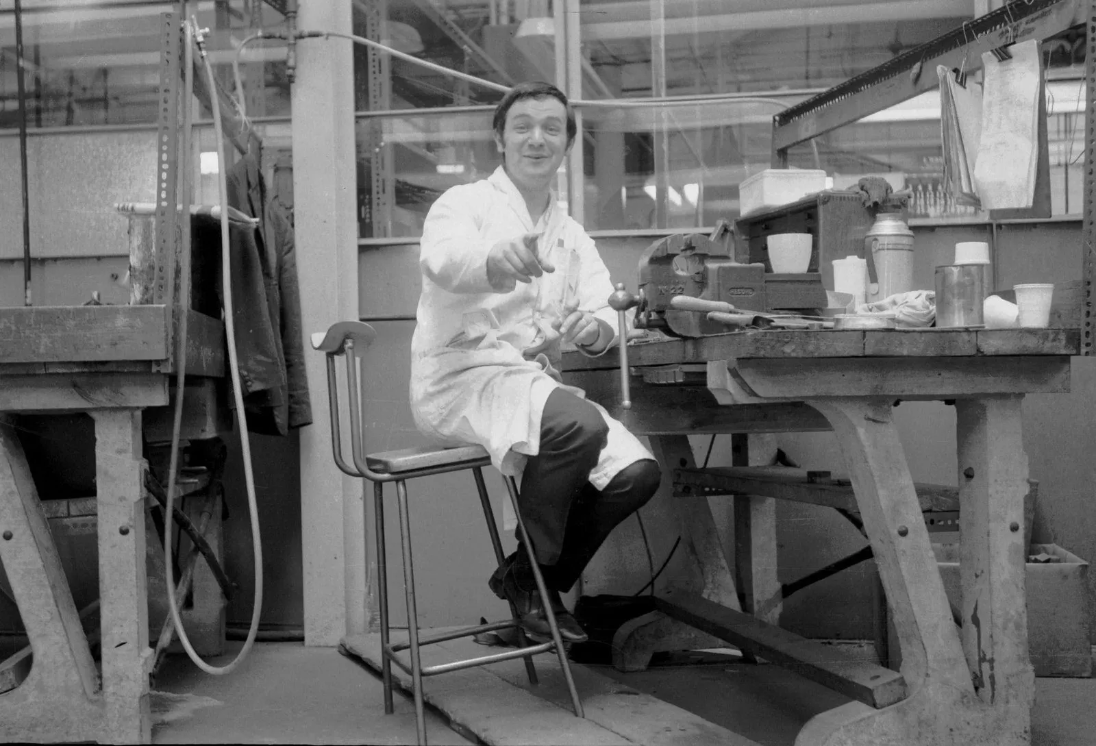 Black-and-white photograph of a Ronson factory worker in a white coat seated at a workbench covered with tools, pointing at the camera and grinning.