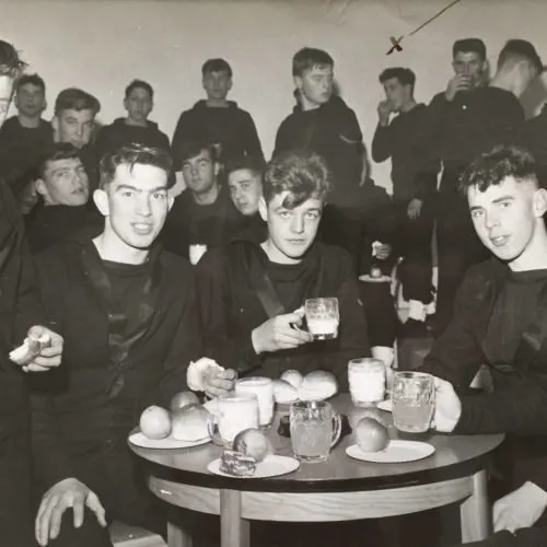 Black-and-white photograph of young sailors in dark jumpers crowded round a table with mugs and fruit, smiling at the camera.
