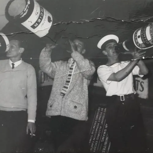 Black-and-white photograph of sailors at a party, one in a white uniform and cap blowing a trumpet.