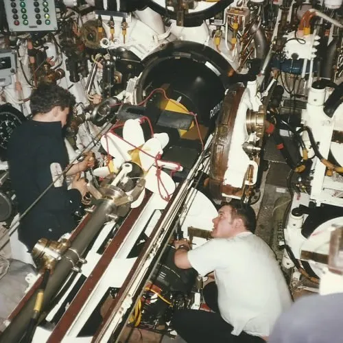 Colour photograph of two crew members working among pipes and machinery in a submarine’s cramped torpedo compartment.