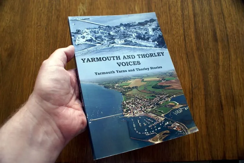 A hand holding a paperback copy of the Yarmouth and Thorley Voices book, with an aerial photograph of Yarmouth on the cover.