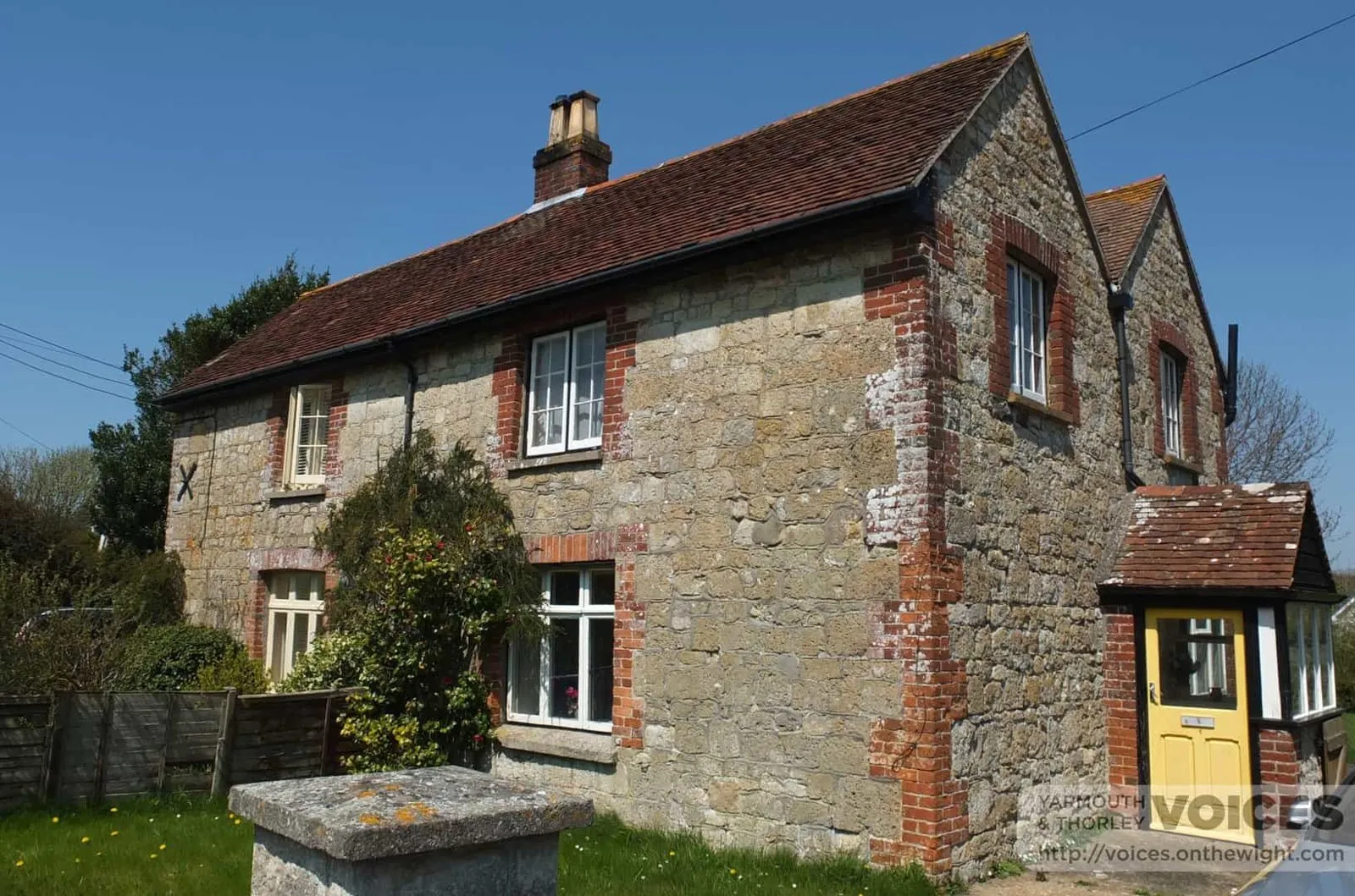 Colour photograph of two joined stone cottages with red-brick window surrounds and a yellow porch door, in spring sunshine.